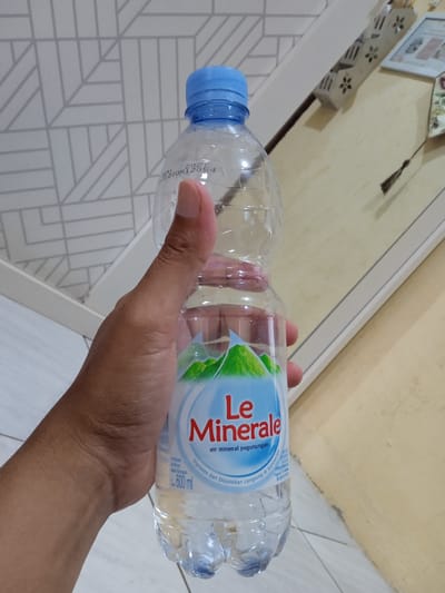 A person's hand holds a clear plastic bottle of "Le Minerale" mineral water. The bottle has a blue cap and the label features green mountains and the product name in red. The text on the label includes "air mineral pegunungan" and "600 ml". The bottle's expiration date on top is NOV 2027. The background includes a geometric patterned wall and a beige surface with decorative elements. The location is Cangkring, Indonesia.