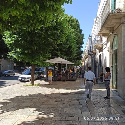 The image depicts a street scene in Ruvo di Puglia, Italy, on July 6, 2026, at 09:51. The perspective is from a sidewalk paved with large, irregular stone blocks, with shadows of trees cast across it.
On the left side of the image, a street lined with parked cars runs parallel to the sidewalk. Notable vehicles include a white Mercedes-Benz sedan with a license plate starting with "GU" parked near a tree, a dark-colored car behind it, and a silver hatchback further down the street. Across the street, buildings with multiple stories and balconies are visible.
The right side of the street features a row of buildings with arched doorways and balconies. A woman in a patterned outfit stands on the sidewalk, facing right and looking at her phone. Further down, a man in a light blue striped shirt and light-colored pants walks away from the camera.
In the middle ground, a group of people are seated at tables outside a cafe, sheltered by a large white umbrella. Several bicycles are parked near the cafe seating area. A woman in a yellow t-shirt and grey shorts stands near the cafe, also looking at her phone.
The environment is characterized by lush green trees lining the street, creating a shaded, outdoor cafe atmosphere. The sky is clear and blue, indicating fair weather. The architecture suggests an urban setting with a historical feel.