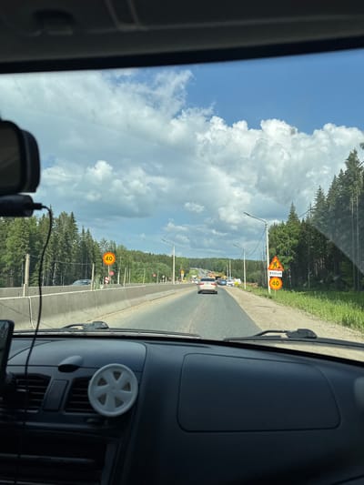 The image captures a view from the driver's seat of a car traveling on a highway with a concrete barrier on the left and a dense forest on the right. The sky is blue with scattered white clouds. Road signs are visible, including a speed limit sign indicating "40" and another sign with a construction worker symbol and "600m." Several cars are on the road ahead. The dashboard of the car is visible in the foreground, with a small, white, flower-shaped object attached to a vent. The road appears to be under construction or repair, with barriers and possibly machinery visible in the distance. The scene suggests a typical highway environment in a rural or semi-rural area, likely in Russia given the context of Nytva.