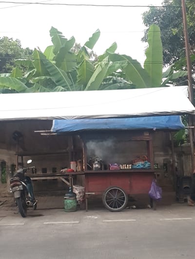 A food stall is set up in front of a concrete building with a white tarp overhead and a blue tarp as a rain cover. A red cart with a spoked wheel is positioned under the tarp, and a motorcycle is parked to the left. Banana leaves are visible in the background. The scene appears to be on a street in Batang, Indonesia. There are no people clearly visible in the frame, but there are items that suggest activity at the stall, such as cooking equipment and a water dispenser. The overall impression is of a humble, functional street-side business.