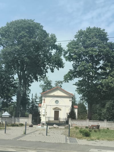 The image shows a view of the Holy Family Church in Łomna, Poland, on a sunny day. The church is a small, pale yellow building with a red-tiled roof and a circular window above the entrance. Two large trees flank the church, their branches extending outward. A low stone wall surrounds the church property, with a black metal fence in front of the entrance. A statue of a saint stands to the left of the entrance. In the foreground, there is a paved area with several bollards and a bench visible in the grassy area to the right. The sky is blue with some scattered clouds.