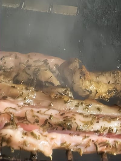 The media file shows a close-up view of meat, likely pork or lamb, being cooked. The meat is arranged in strips, with visible herbs and seasoning, possibly rosemary, sprinkled on top. The cooking method appears to be roasting or grilling, as evidenced by the char marks on some pieces and the overall texture. There is a smoky or steamy atmosphere surrounding the meat, suggesting it is actively being cooked. The background is dark and somewhat out of focus, providing no clear environmental details. The lighting is dim, highlighting the textures and colors of the meat. No people, text, time of day, or weather cues are discernible. The context provided indicates the location is Zlataritsa, Bulgaria.
