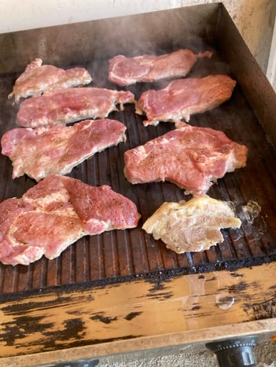 This image captures a close-up, high-angle view of multiple cuts of meat grilling on a ribbed, flat-top griddle in Raduil, Bulgaria. Eight pieces of raw or partially cooked, seasoned pork steaks, primarily pinkish-red with visible white fat marbling, are distributed across the dark, well-used metal surface of the grill. Wisps of white smoke or steam are rising from the cooking meat, particularly in the upper half of the frame. Rendered fat and juices accumulate in the grill's channels and along its front edge, where one smaller, lighter, and more cooked piece of meat/fat is visible. The grill rests on a light brown surface, possibly wood or composite material, with a partially visible black control knob in the bottom right. A light-colored, possibly concrete or stone wall, serves as a background directly behind the grill. No individuals are visible in the scene.