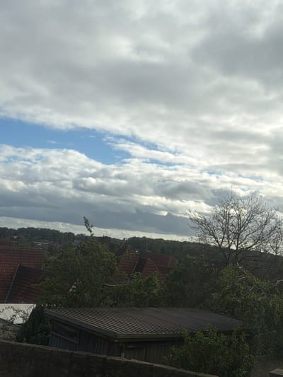 The image shows a view of the sky and a portion of Bad Bentheim, Germany. The sky is filled with clouds in shades of gray and white, with patches of blue sky visible. A leafless tree is silhouetted against the sky. Below the tree, the tops of houses with red tile roofs are visible. A dark shed or building with corrugated metal siding is in the foreground, near what appears to be a stone wall. The overall scene is a combination of natural and built elements in the Bad Bentheim landscape.