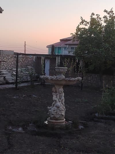 The image depicts a garden scene in Ignatievo, Bulgaria, during the soft light of either dawn or dusk. A decorative fountain, sculpted with figures of cherubs or putti, stands prominently in the foreground. The fountain appears to be made of stone or concrete and has multiple tiers. The surrounding ground is dark, uncultivated soil with some sparse vegetation.
In the mid-ground, a stone wall forms a boundary. Piles of flat, rectangular objects, possibly tiles or stone slabs, are stacked against this wall. A dark metal structure, likely a frame or support, is also visible near the wall. Further back, a house with a red-tiled roof and white railings on a balcony is partially obscured by a leafy green tree. Electrical wires and a utility pole are visible in the upper left portion of the image, indicating the presence of infrastructure. The sky displays a gradient of colors, transitioning from a pale orange near the horizon to a lighter hue towards the top. There are no people visible in the image, and the overall atmosphere suggests a quiet, perhaps unmaintained, outdoor space.