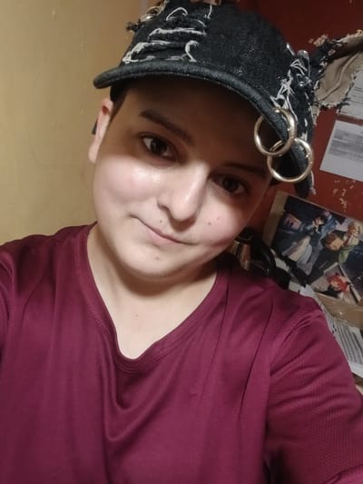 The image is a close-up, selfie-style shot of a person wearing a dark, distressed baseball cap with silver rings and white stitching. The person has short dark hair, visible on the sides of the cap. They are wearing a maroon t-shirt. The background is a wall with a textured yellow surface on the left and a darker, reddish-brown surface on the right, adorned with papers and what appears to be an anime-style print. The overall lighting suggests an indoor environment. The image does not contain any information that would allow for the identification of its location within Chutove, Ukraine.