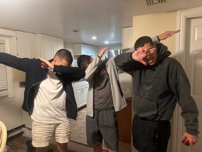Three young adult males are posing for a photo inside a residence, likely in Ocean City, Maryland, USA. The individuals are making a "dab" gesture, with one person's face obscured by their hand. They are dressed casually in t-shirts, hoodies, and shorts. The background appears to be a kitchen or living area with white cabinets, a countertop, and a doorway. The lighting suggests it is daytime or early evening.