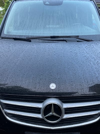 A black Mercedes-Benz vehicle is depicted in a close-up, frontal view, located in Куликово, Russia. The vehicle's windshield, hood, and front grille are densely covered with water droplets and streaks, indicating rain. Two black windshield wipers are positioned at the base of the windshield. The black hood features a circular silver Mercedes-Benz badge. A large, central silver Mercedes-Benz three-pointed star emblem is prominent on the multi-slatted front grille. Blurred green foliage is visible through the upper portion of the wet windshield. No individuals are present.