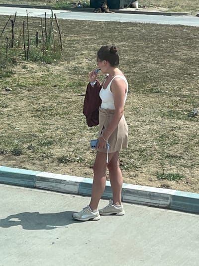 A young woman with her hair in a bun stands on a sidewalk, facing right. She is wearing a white tank top, a tan skirt, and white sneakers. She is holding a blue object, possibly a phone, in her right hand and a dark bag in her left. A faint shadow of the woman is cast on the ground in front of her. The background consists of dry grass and some wooden poles. In the distance, a road and some structures are visible. The scene appears to be in a semi-urban or rural area. The location is identified as Ovidiu, Romania.