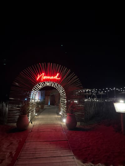 This image depicts the entrance to a establishment called "Nomad" in Torreilles, France, at night. A pathway made of weathered wooden planks leads through a semicircular archway constructed from numerous thin wooden poles. The archway is adorned with a bright red neon sign spelling out "Nomad" in cursive. On either side of the pathway, large, terracotta-colored urns hold palm-like plants. The ground on the sides of the pathway is sand, illuminated by red ambient lighting. Beyond the archway, more wooden fencing and a line of string lights are visible, suggesting an outdoor seating area or beachside establishment. The sky is dark and clear, with a few faint lights visible in the distance. The overall scene conveys a festive and inviting atmosphere, likely a restaurant, bar, or beach club.