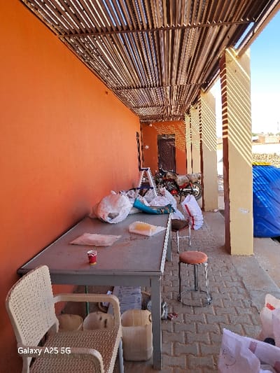 The image shows a side view of a building in Karima, Sudan (18.54, 31.831). It is a building with a red brick exterior, a roof made of wooden slats, and an uncovered patio area. The patio has a concrete floor, a long table with plastic bags and other objects scattered on its surface, two chairs, and two stools. One of the chairs is a white plastic chair. One of the stools is a wooden stool painted red. There is a red cup on the table. There is a blue plastic barrel on the far right of the image. The wall of the building is painted a bright orange. There is a ladder and some motorcycles in the background of the image. It appears to be daytime, with the sun shining brightly.