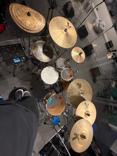 The image presents an overhead view of a drum set and accompanying musical equipment, likely situated in a recording studio or practice room in Shemyakino, Russia. The perspective is from above, offering a glimpse of the setup from the drummer's position.
The central focus is an acoustic drum kit with a dark wood finish, comprising a snare drum, tom-toms, and a kick drum. The white snare drumhead clearly displays the "REMO" brand name. Numerous golden cymbals are arranged around the kit; several prominently feature the "K" logo and the text "K CUSTOM DARK," identifying them as Zildjian K Custom Dark series cymbals. A pair of stacked cymbals on the far left also shows a distinct circular logo.
To the right, the lower legs and feet of a person wearing dark pants and dark Crocs shoes are visible, indicating the drummer is seated at the kit. An electronic sampling pad, identified as a Roland "SPD-SX," with glowing red and green pads, is positioned on the floor to the left of the drum pedals.
The drum kit is partially enclosed by clear acrylic sound baffles, suggesting sound isolation or control. Several microphones on stands are placed around the kit, along with various black studio monitors or speakers on the carpeted floor. In the background, a grey bean bag chair, cables, and other equipment are visible. The scene depicts a well-equipped indoor musical environment, likely illuminated by artificial light.