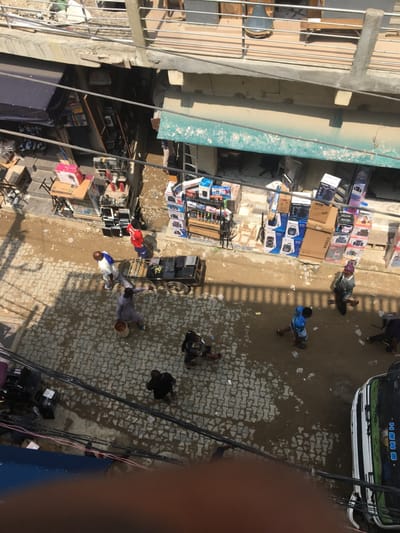 The image presents an elevated view of a street in Ojo, Nigeria, under bright daylight with distinct shadows. The street surface transitions from a patterned grey paver section on the left to a dirt and mud section on the right, both showing evidence of moisture and puddles. Numerous black electrical or communication cables crisscross overhead.
On the left, several vendor stalls operate under dark awnings, displaying electronics and various components on tables and ground arrangements. On the right, a building with a light green awning features extensive displays of stacked, boxed electronic items, including speakers and appliances, extending onto the street level. An upper story balcony with concrete railings and discernible objects is visible above these structures.
Approximately ten individuals are present at street level, engaged in various activities. In the center-left, two men interact near a two-wheeled handcart loaded with dark, rectangular objects, likely electronic devices. One man wears a white and blue garment, the other a light brown traditional outfit. A person in a red shirt stands near a stall on the left. On the right, a man in a blue shirt walks on the dirt section, where plastic litter is scattered. A white and blue bus or commercial vehicle is partially visible, parked on the far right of the dirt section. People are observed walking and standing, indicating general street commerce and pedestrian traffic.