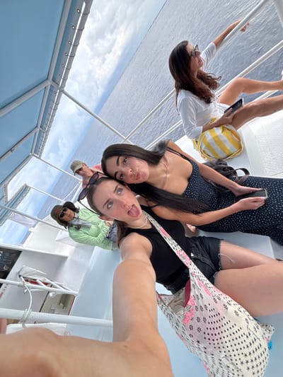 The image depicts five individuals on a white passenger boat on open water off West Bay, Cayman Islands. The scene is a selfie, with the primary subject, a young adult female with light brown hair, visible from the chest down in the foreground. She wears a black tank top and dark shorts, with a white, pink, and black patterned tote bag over her left shoulder. Her expression shows her tongue slightly extended and eyes gazing upwards; sunglasses are perched on her head.
Immediately to her right, a second young adult female with long dark hair, wearing a dark blue patterned dress, holds a smartphone in her left hand and displays pouting lips towards the camera. Behind them, an older adult female in a light green long-sleeved shirt and a wide-brimmed hat is seated on the left, and an older adult male in a light-colored polo shirt and baseball cap is partially visible behind her; both wear sunglasses. On the right side of the boat, a third adult female with long dark hair, wearing a white long-sleeved shirt, is seated, holding a smartphone, and observing the water with her right arm resting on the boat's railing. A yellow and white striped bag rests beside her.
The boat features white deck surfaces, white railings, and a light blue canopy structure overhead. The surrounding environment shows calm blue ocean water extending to a distant horizon under a partly cloudy sky.