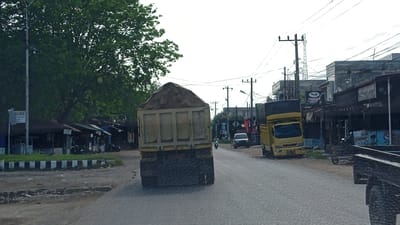 The image depicts a street scene in Uteungeulinggang, Indonesia. A large, yellow dump truck loaded with sand is the primary subject in the foreground, moving away from the camera. To its left, a street sign with the text "MACHMUR" is visible near a row of small shops or stalls. Further down the road, another yellow truck is parked, and a white car is also present. The environment includes a paved road, a sidewalk with black and white barriers, a large tree on the left, and buildings lining the right side of the street. Several utility poles with electrical wires are scattered throughout the scene. The sky is overcast, suggesting a cloudy day.