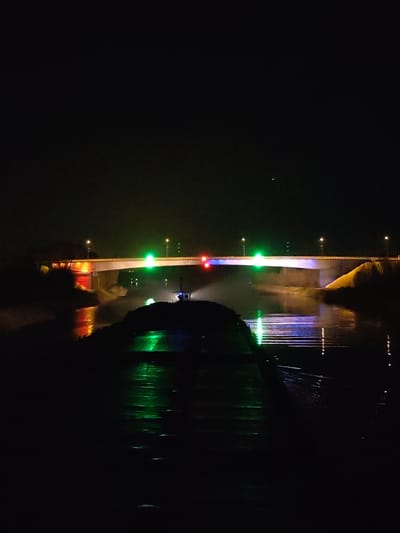 At night, the image depicts a barge on the Oka River near the city of Podosinki, Russia. The barge is dark, with a green light illuminating its side and reflecting on the water. In the background, a bridge spans the river, lit with green and red lights. Streetlights line the bridge. The water reflects the bridge's lights. The sky is dark. A small light is visible in the sky. Vegetation is visible on both sides of the river.