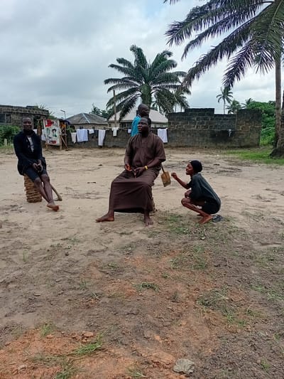 In the city of Ipota, Nigeria, four individuals are present in an outdoor, sandy area. The scene is characterized by a rough, dirt ground with sparse patches of grass and some scattered debris. A low, dark brick wall forms a partial backdrop, with laundry hanging on a clothesline in front of it. Beyond the wall, residential buildings and palm trees are visible under an overcast sky.
On the left, a young person is seated on a cylindrical object, possibly a log or a cut tree trunk. This individual wears a black jacket, shorts, and white socks, with their bare feet on the ground.
To the right of the first individual, an adult male is seated on the ground, wearing a loose, brown robe-like garment. He appears to be in conversation.
In the center, another adult male stands, partially obscured by the seated individuals. He is wearing a blue garment.
In the foreground on the right, a young person is crouched down, wearing a dark t-shirt and shorts, and a black head covering. They are holding a small, light-colored bag and appear to be actively engaged in conversation or interacting with the seated man. Their bare feet are visible. A pair of sandals lies on the ground near them.
