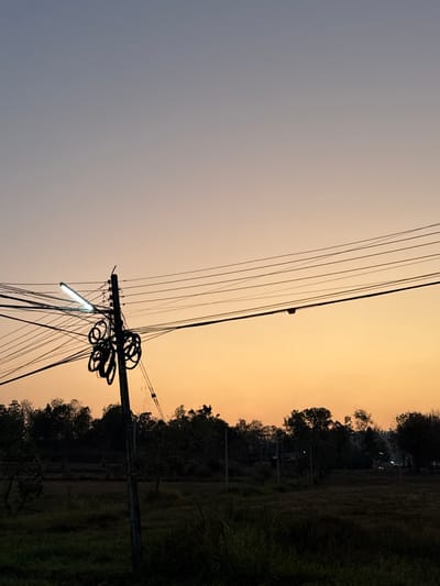 The scene is located in Ban Huai Sai, Thailand, at dusk or dawn. The sky displays a gradient, transitioning from a light blue-grey at the zenith to an orange-yellow hue near the horizon. In the left-center foreground, a dark utility pole stands vertically. Attached to this pole is a rectangular, illuminated fluorescent light fixture emitting a bright white-blue light. Numerous black electrical and communication wires extend horizontally across the frame, originating from and connected to the pole, with several bundles of coiled black wires also affixed to the pole. The midground and background consist of a dark silhouette of dense trees and foliage, appearing as an irregular horizontal band below the sky. Beneath the tree line, the ground is covered with dark, low-lying vegetation or grass. Faint, distant light sources, possibly from buildings or vehicles, are visible through the trees on the far right. No human figures are present.