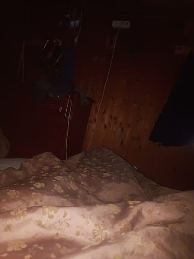 The image depicts a dimly lit interior of what appears to be a bedroom or attic space, likely located in Vysokovo, Russia. The walls and ceiling are constructed of dark, rustic wood paneling. In the background, a red dresser or cabinet is visible, with some items, possibly electronics, perched on top. A white electrical extension cord snakes down from the ceiling, plugging into a multi-socket adapter.

In the foreground, a bed is covered with a patterned duvet or blanket. The fabric is a light pink or peach color with a repeating floral motif in a darker pink or brown hue. The bedding is rumpled, suggesting recent use or disarray. To the right, a dark, amorphous shape is draped against the wooden wall, possibly a piece of clothing or fabric. The overall atmosphere is one of subdued lighting and a somewhat cluttered domestic environment. No people are visible in the frame.