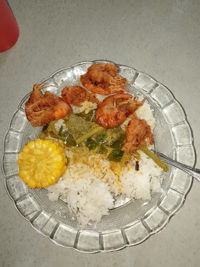 This media file captures a close-up, overhead view of a plate of food. The main subjects are several pieces of fried shrimp, a mound of white rice, a portion of green vegetables in a sauce, and a slice of corn on the cob. The plate is made of clear, textured glass with a scalloped edge. A metal fork is partially visible on the right side of the plate, resting on the food.

The food appears to be a savory meal, likely intended for consumption. There are no people visible in the frame, nor is there any indication of a specific time of day or weather conditions. The background is a neutral, textured surface, possibly a table or countertop. Given the provided location context of Meunasah Kulam, Indonesia, the meal could be a common local dish. The image is focused on the food itself, suggesting it's meant to showcase the meal. There is no visible text in the image.