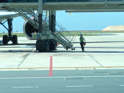 An airplane is parked on an airport tarmac. A set of mobile stairs is connected to the airplane's fuselage. A person in a high-visibility vest and blue cap is walking away from the stairs, holding a red folder. The background shows a clear blue sky and a sandy hill. The scene appears to be at Cam Ranh International Airport in Cam Ranh, Vietnam, based on the overall environment and typical airport infrastructure.