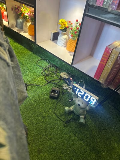 The image depicts an indoor scene within a commercial or display area in Naze, Nigeria. The floor is covered with artificial green turf. Along the background, a series of vertically arranged white display shelves are visible, each internally illuminated. The shelves on the left contain multiple decorative vases of varying shapes and colors (red, light blue, white, orange) holding artificial flowers. A small white rectangular object is also present on one of these shelves. On the right-side shelves, additional vases with artificial yellow sunflowers and red berry-like flowers are displayed. Below these, a stack of books, wrapped in clear plastic, features visible titles such as "Art," "VERSACE," and "WATCHES."
On the artificial turf floor, a tangled arrangement of black electrical cables, a black power strip with multiple occupied outlets, and a black battery charger are present. Positioned on the turf, centrally to the lower right, is a light green/teal figurine, resembling a KAWS companion, which holds an illuminated white digital clock displaying "12:09" in bright blue LED digits. A white USB cable connects to the clock. The lower-left foreground of the image shows the partial view of a person's left leg, clad in light blue denim jeans, suggesting a close, low-angle perspective. The environment is uniformly lit by artificial light sources originating from within the display units and the digital clock.