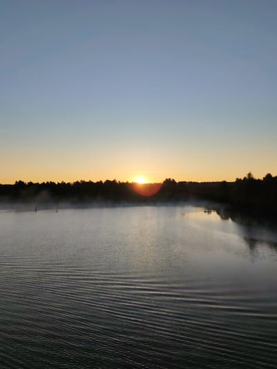 This is a photograph of a sunrise over a lake in Yalosar', Russia. The sun is low on the horizon, casting a warm, orange glow across the sky and reflecting off the calm water. A light mist hangs over the lake, creating a serene and peaceful atmosphere. The shoreline is lined with trees, their dark silhouettes providing a stark contrast to the bright sky. There are no people or animals visible in the image, and the scene appears undisturbed. The photograph captures a moment of natural beauty and tranquility.