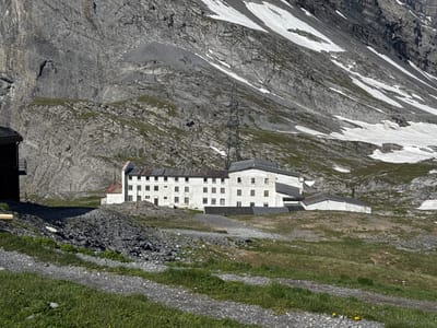 An outdoor, medium-distance, eye-level, horizontal, clear, daylight shot shows a series of old, white, multi-story buildings with weathered, rusty-colored roofs, nestled at the base of a steep, rocky mountain face with patches of snow. The buildings appear to be an abandoned hotel or similar complex. A tall, metal power transmission tower stands behind the buildings. The foreground consists of a gravel path winding through green grass and a pile of dark, sharp rocks. The environment is mountainous and rugged. There are no people visible in the image. This scene is likely the historic Grand Hotel de la Poste or a related structure in the Gemmi Pass area above Leukerbad, Switzerland, known for its former status as a mountain resort and its historical significance.