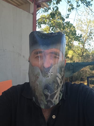 The image shows a person wearing a black collared shirt and a custom-made mask that covers their face and head. The mask appears to be constructed from a dark, possibly plastic material, with a clear face shield. The face shield has a camouflage pattern spray-painted onto it in patches of green and grey, and is secured to the rest of the mask with visible fasteners. The person's eyes are visible through the clear portion of the face shield. The background suggests an outdoor setting, possibly a rural or semi-rural area. There is a light-colored concrete wall to the left, and trees with green foliage and a blue sky are visible in the background to the right. A structure with a corrugated roof and red supports is partially visible at the top of the image. The image is a close-up, likely a selfie, focusing on the person's face and upper body. No specific landmarks in Çatalca, Turkey, are identifiable.