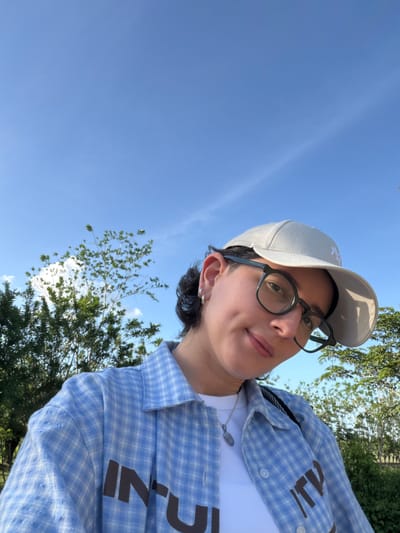 A low-angle outdoor shot shows a person with light skin, wearing glasses and a light-colored baseball cap, facing the camera with a slight smile. They are wearing a light blue and white plaid shirt over a white t-shirt. A silver necklace with a pendant is visible. Parts of a tree and foliage are in the lower right corner. The sky is blue and largely cloudless in the upper left portion. It's impossible to determine if the photo was taken in Mata De Caña, Colombia from visual features.