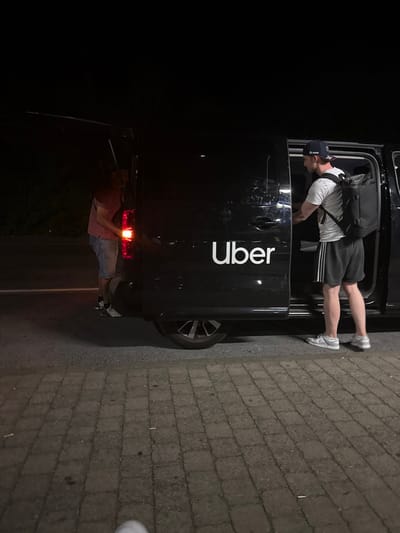 A man is reaching into the back of a black Uber vehicle. He is wearing a white shirt, dark shorts, a baseball cap, and a backpack. The vehicle is parked on a brick surface, and the passenger side door is open. The rear door is also ajar, and another person is possibly visible in the back. The scene is poorly lit, with the rear light illuminated.