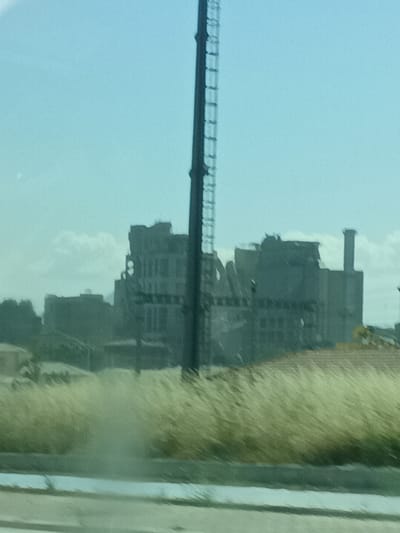 The image depicts a somewhat hazy view of a damaged or partially demolished industrial building complex in Konya, Turkey. In the foreground, there is a strip of dry, tall grass, suggesting an arid or semi-arid environment. Behind the grass, the industrial structures dominate the scene. These buildings appear weathered and incomplete, with some sections showing signs of ruin or deconstruction. One prominent feature is a tall, narrow smokestack or tower on the right side of the frame, with a narrower, lighter-colored chimney-like structure next to it. A very tall, dark, lattice-like structure, possibly a radio tower or a part of the industrial complex, runs vertically along the right edge of the image, partially obscuring the sky and buildings.
The sky is a pale, washed-out blue with a few scattered, wispy clouds, indicating it could be daytime, likely late morning or afternoon given the brightness and lack of strong shadows. The overall atmosphere of the image is somewhat desolate and industrial. There are no people visible in the frame. The scene appears to be taken from a moving vehicle, given the slight blur and the perspective. No discernible text is visible in the image.