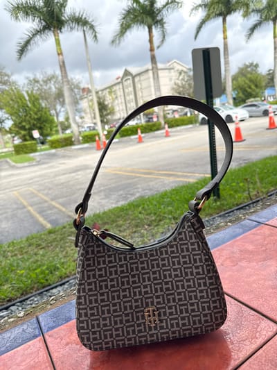 A Tommy Hilfiger purse, patterned with gray and dark brown "TH" logos, sits on multi-colored square tiles, next to grass. The purse handle is black with gold hardware. The background depicts a street lined with palm trees, parked cars, and orange cones. A white building is partially visible in the distance, along with an obscured street sign. The weather appears overcast.