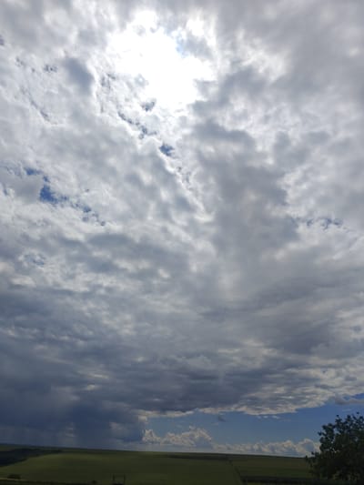 The image captures a wide-angle view of a sky filled with varied cloud formations over a rural landscape. In the upper portion of the image, the sun is partially obscured by bright, white clouds, creating a luminous effect. The clouds transition to darker, more ominous gray tones as they descend towards the horizon.

The lower part of the image reveals a green field, suggestive of agricultural land, stretching across the scene. A dark line, possibly a tree line or a distant embankment, delineates the edge of the field. In the far right of the frame, a portion of a leafy green tree is visible, its dark silhouette contrasting with the lighter sky. The overall atmosphere appears to be one of an overcast or transitional weather day. The location is identified as Colonița, Moldova.