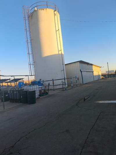 A large, white, cylindrical silo stands prominently in an outdoor industrial setting. A metal ladder with safety cages runs up the side of the silo. To the right of the silo is a metal-clad building with large sliding doors. In the foreground and to the left of the silo, several green and white drums are stacked on pallets. Blue barrels and other containers are also visible. A chain-link fence with blue and white posts runs along the foreground. The scene appears to be a storage or processing facility.
The sun is low in the sky, casting long shadows and illuminating the silo with a warm, golden light, suggesting either early morning or late afternoon. The sky is clear and blue. The ground is paved with asphalt, showing cracks and a puddle of water. In the background, there are some solar panels and other structures, indicating an open, potentially rural or semi-industrial area. There is no visible activity or people in the image.
A small white sign is affixed to the silo, but the text is too small to be legible. The overall impression is one of a quiet, functional industrial space bathed in the soft light of either dawn or dusk.