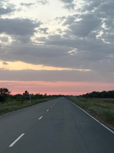 The image depicts an asphalt road stretching into the distance under a cloudy sky at dusk. The sky displays hues of orange and pink, indicative of a sunset or sunrise, with darker clouds in the upper portion. The road has white lane markings. On either side of the road are grassy fields and trees, with a signpost visible in the left mid-ground. The scene appears to be in a rural or semi-rural setting, potentially the outskirts of Пугачёв ауылдық округі, Kazakhstan, given the context. There are no people or animals visible in the image.