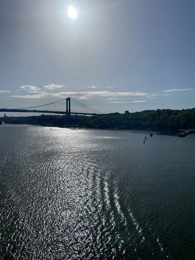 The image captures a view of the Göta älv river in Gothenburg, Sweden, under a bright, sunny sky. A large suspension bridge, the Älvsborg Bridge, spans the river, with its distinctive towers and cables clearly visible. The bridge carries vehicles and is a prominent landmark. On the far bank, a densely wooded area with scattered buildings creates a green, natural backdrop to the urban infrastructure. The river's surface is choppy, with sunlight reflecting off the waves, creating a shimmering effect. Several buoys are visible in the water, indicating navigation markers. The overall scene depicts a dynamic waterfront with a blend of natural and man-made elements.