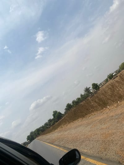 The scene, located in Mayo, Nigeria, is a roadside view captured from a moving vehicle. The lower left quadrant of the image shows a portion of a dark-colored vehicle, including its side mirror. The paved road, visible from the bottom center extending into the mid-distance on an upward incline, features a solid double yellow line marking its edge. To the right of the road, the immediate shoulder consists of dry, reddish-brown soil, transitioning into a strip of tall, desiccated brown grass and sparse low-lying vegetation. Beyond this dry vegetation, a line of green trees, including several palm trees, runs parallel to the road. Further in the background, faint structures or buildings are partially visible among the distant trees. The sky occupies the upper two-thirds of the image, displaying a light blue hue with scattered white cumulus and wispy cirrus clouds. No individuals or specific interactions are discernible in the frame.