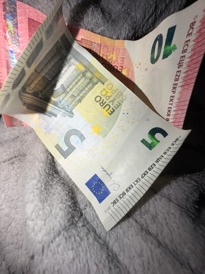 The image displays two euro banknotes, a 5 euro note and a 10 euro note, partially unfurled and resting on a textured gray surface, possibly a blanket or carpet. The banknotes are the primary subjects. The 5 euro note is more prominent, showing the number "5" in a metallic green ink and the European Union flag. The text "BCE ECB EЦБ EZB EKP EKT EKB BCE EBC 2013" is visible along the top edge of the note. The 10 euro note, partially obscured, shows the number "10" in a similar metallic green ink and the text "BCE ECB EЦБ EZB EKP EKT EKB B". The overall lighting suggests an indoor setting, possibly taken in the evening or with artificial light. The focus is on the details of the banknotes, highlighting their design elements and markings. There is no indication of people, specific activities, or a particular time of day or weather. The location context is Dorsten, Germany, but this is not visually discernible from the image itself. The scene is simple and focuses on the currency.