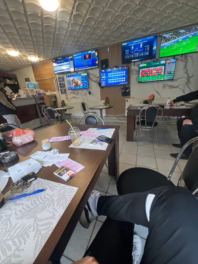 This image shows the interior of what appears to be a betting shop, possibly in the city of Batoshevo, Bulgaria, given the presence of Bulgarian language text on the screens. The room contains multiple televisions mounted on a wall, displaying various sports events and betting odds.
Several individuals are present, with at least two visible seated at tables. These people appear to be patrons or employees of the establishment.
The tables are cluttered with betting slips, receipts, keys, and other miscellaneous items, including a pen and an empty plastic cup.