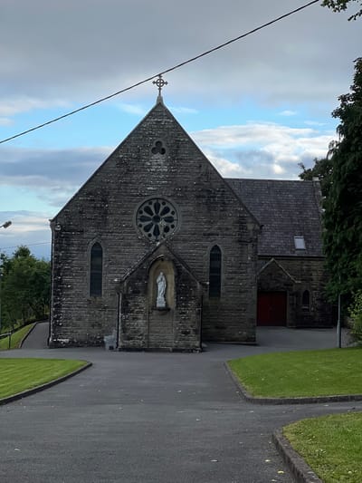 This is a photograph of a stone church, taken from a low angle looking up towards the sky. The church has a peaked roof and a large circular window with intricate stonework. A statue of a robed figure, likely a religious icon, is set in an alcove to the left of the circular window. Above the main entrance, a small peaked gable features a red door. The stonework of the church is weathered, with patches of moss and lichen.
To the left of the church, a paved pathway curves around a manicured lawn. The sky above is dramatic, with a mix of bright blue patches and heavy grey clouds, suggesting a changeable weather day. A thin black wire runs diagonally across the upper right portion of the image. A cross-shaped finial is visible at the apex of the church's roof, silhouetted against the sky. Trees and foliage are visible at the bottom of the frame, framing the lower part of the church. The scene is tranquil, with no people visible. The lighting suggests it is either late afternoon or early morning. The presence of the cross and religious statue firmly places the setting as a place of worship. Based on the location context provided, this is likely Mountcharles, Ireland.