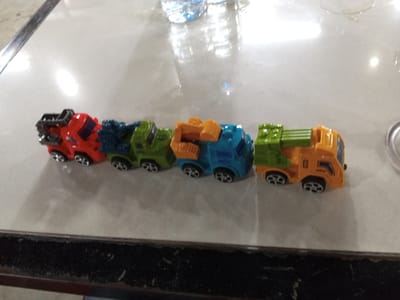 The image shows a line of five toy trucks on a reflective surface, likely a table. From left to right, the trucks are red, blue, green, blue with an orange scoop, and orange with a green back. The trucks appear to be part of a construction or garbage collection toy set, with various attachments like scoops and bins. The background is blurred, but suggests an indoor setting with some reflections indicating light sources from above. Two partially visible plastic water bottles are in the upper background. The overall lighting is bright, suggesting daytime. There are no people visible. The surface they are on is light-colored and shows some wear and tear, particularly a dark border at the bottom of the frame that looks like a textured edge. The toys are the main focus, arranged in a neat line.
