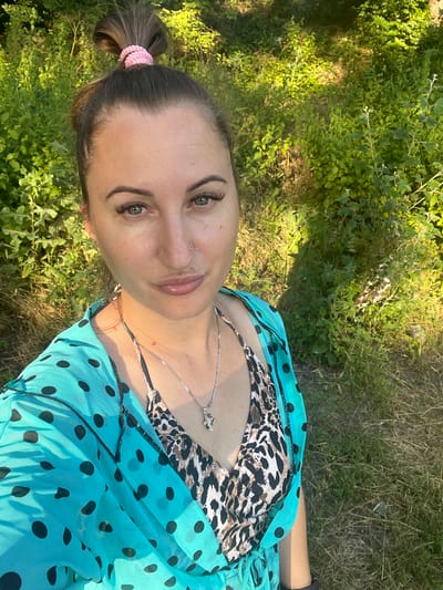 A selfie shows a woman with her hair in a bun with a pink scrunchie, standing outdoors. She is wearing a turquoise polka dot cover-up over a leopard print bikini top. She has on a silver necklace with a charm. The background consists of lush green foliage and dry grass, suggesting a natural, possibly rural, environment. The lighting indicates it is daytime, likely with sunlight filtering through the trees. The location is identified as Zarichne, Ukraine.