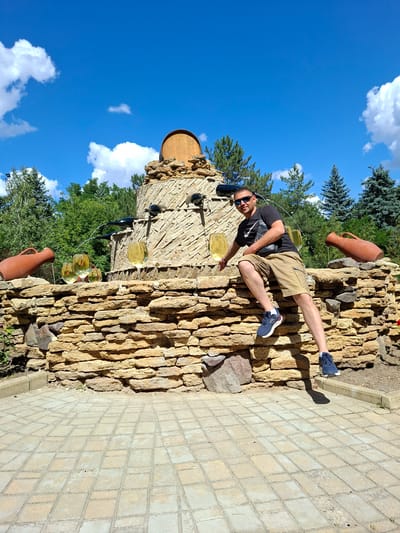 A man is posing for a photo in front of a wine-themed fountain at a winery in Milestii Mici, Moldova. The fountain is made of stone and features wine bottles and large wine glasses. The man is wearing sunglasses, a black t-shirt, and tan shorts, and is sitting on the edge of the fountain with his legs extended. The sky is blue with white clouds, and there are trees in the background. The ground in front of the fountain is paved with square tiles.