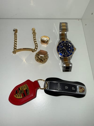A top-down, medium shot shows a Rolex watch, two gold rings, a gold bracelet, and a Porsche car key with a red fob on a white surface. The Rolex is a two-tone stainless steel and gold Submariner with a blue dial and bezel. The rings are gold; one is a signet ring with intricate patterns, and the other is a square-shaped ring with a small diamond or stone. The gold bracelet has a mix of curb chain and bar links. The Porsche car key is black, with a silver accent piece. The red key fob has the Porsche crest emblazoned on it. The setting is indoors, suggesting the objects are placed on a shelf or table. There is no indication this photograph was taken in the city of Bredene, Belgium.