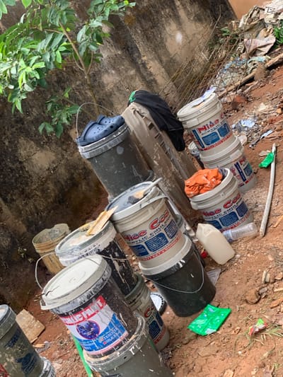 The image depicts an outdoor scene in Amaeshi, Nigeria, characterized by an accumulation of discarded and used materials against a weathered concrete wall. The foreground and midground are dominated by numerous plastic paint buckets, varying in color from white to dark grey/black, many exhibiting splatters and residue of paint. Several white buckets are stacked, prominently displaying labels such as "Nide Acrylic Emulsion" and "Nice Coat Acrylic Emulsion." A pair of blue Crocs brand sandals rests on top of a dark grey bucket. An orange fabric item, possibly a rag or clothing, is visible inside one white bucket, while a black fabric item is draped over a light-colored object, potentially another bucket or a piece of furniture, further back. The reddish-brown soil ground contains scattered debris including a light beige plastic jerrycan, a long white pole, and a green plastic wrapper. In the background, a small tree with green leaves grows near the concrete wall, which shows signs of age and discoloration. Further to the right, behind the stacked buckets, exposed metal rebar or mesh is visible amidst general refuse and torn packaging. No individuals are present in the scene.