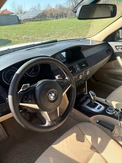 The image displays the interior of a BMW vehicle, specifically the driver's perspective, looking forward through the windshield towards a rural outdoor scene in Daskal-Atanasovo, Bulgaria.
The vehicle's interior features a black leather-wrapped steering wheel with a central BMW logo, silver paddle shifters, and integrated control buttons. The instrument cluster, showing a speedometer and tachometer, is visible through the steering wheel. The dashboard is multi-toned, incorporating dark and beige sections, with a dark, possibly suede-textured panel on the passenger side. A rearview mirror is mounted on the windshield, from which a pink and white striped air freshener is suspended. The center console includes an automatic gear selector, climate controls, media controls, and an iDrive controller. The driver's seat and parts of the passenger seat are upholstered in beige leather. Dark, textured floor mats cover the footwell.
The exterior view through the windshield shows a brightly lit, green grassy area. To the left, a building with a reddish-brown tiled roof is visible. In the center-right, a ground-mounted solar panel array is prominent. The background features several bare-branched trees and additional structures with reddish roofs, under a clear blue sky. No individuals are present in the scene.