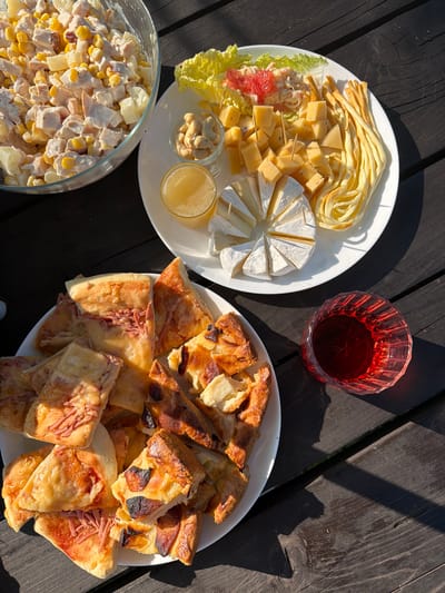 A table setting with food is pictured outdoors, with a wooden table surface that appears weathered and dark. In the top left, a large glass bowl is filled with a salad containing chicken, corn, and possibly pineapple. To the right, a white plate holds a cheese platter with various cheeses, lettuce, grapefruit segments, a small glass of honey, and a cluster of cashews. Below this, another white plate is laden with slices of pizza and some other baked goods, possibly quiche or savory pie. A faceted glass filled with a dark red beverage, likely wine, is situated to the right of the pizza plate. The scene is lit by natural sunlight, casting shadows across the table. There are no people visible in the image. The setting appears to be a casual outdoor meal. The location is specified as Kuliki, Russia.