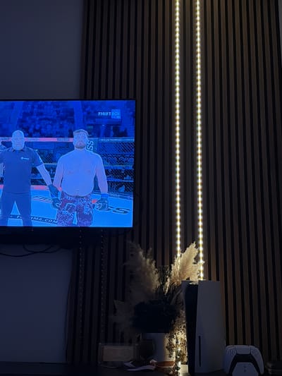 The media file depicts an indoor scene, likely a living room or entertainment area, centered around a television displaying a combat sports event. The TV is mounted on a wall featuring horizontal wooden slat paneling. Two bright LED strip lights run horizontally across the slatted wall above the television, providing artificial illumination.
On the television screen, a mixed martial arts (MMA) or boxing match is in progress. Two men are prominently featured: a shirtless fighter with a beard, wearing patterned shorts and combat gloves, and below him, a referee or official dressed in dark clothing, wearing a black glove on one hand. The background shows elements of a fighting cage and an audience. Visible text on the screen includes "FIGHT BOX", "HD", "TOTALBYGGEN", "SS", "MMA.COM", "WILEA NORDIC", "RANSLAA", "taio out", and "FVB".
To the right of the television, a white Sony PlayStation 5 console stands vertically on a surface, accompanied by a white PlayStation DualSense controller. Behind the console and controller, decorative elements such as dried pampas grass and darker, dried floral arrangements are visible, along with a small, light-colored decorative item. The overall lighting suggests it is evening or night, with the primary light sources being the LED strips and the TV screen. Cables are visible behind the television setup.
