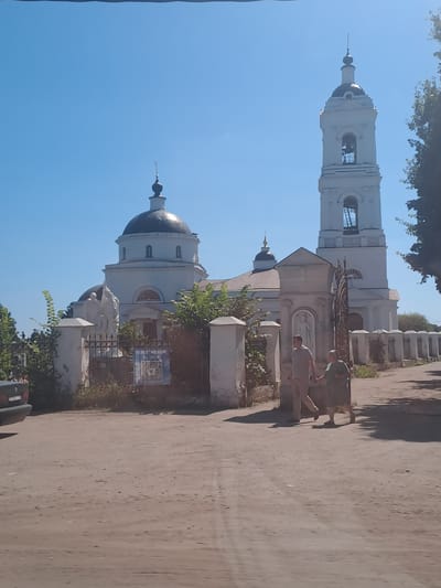 The image shows a white church with a dark dome and a tall bell tower, characteristic of Russian Orthodox architecture, situated in Bogoslovo, Russia. A wrought-iron gate and a section of a white fence with rounded posts are visible in the foreground. Two individuals, a man and a woman, are walking hand-in-hand towards the church entrance. The man is wearing a light-colored polo shirt and light-colored pants, while the woman is dressed in a dark green dress. They are walking on a dirt road leading up to the church. A blue sign with text and an image is affixed to the fence to the left of the gate. Lush green foliage surrounds the church and fence area. The sky is clear and blue, indicating a sunny day.